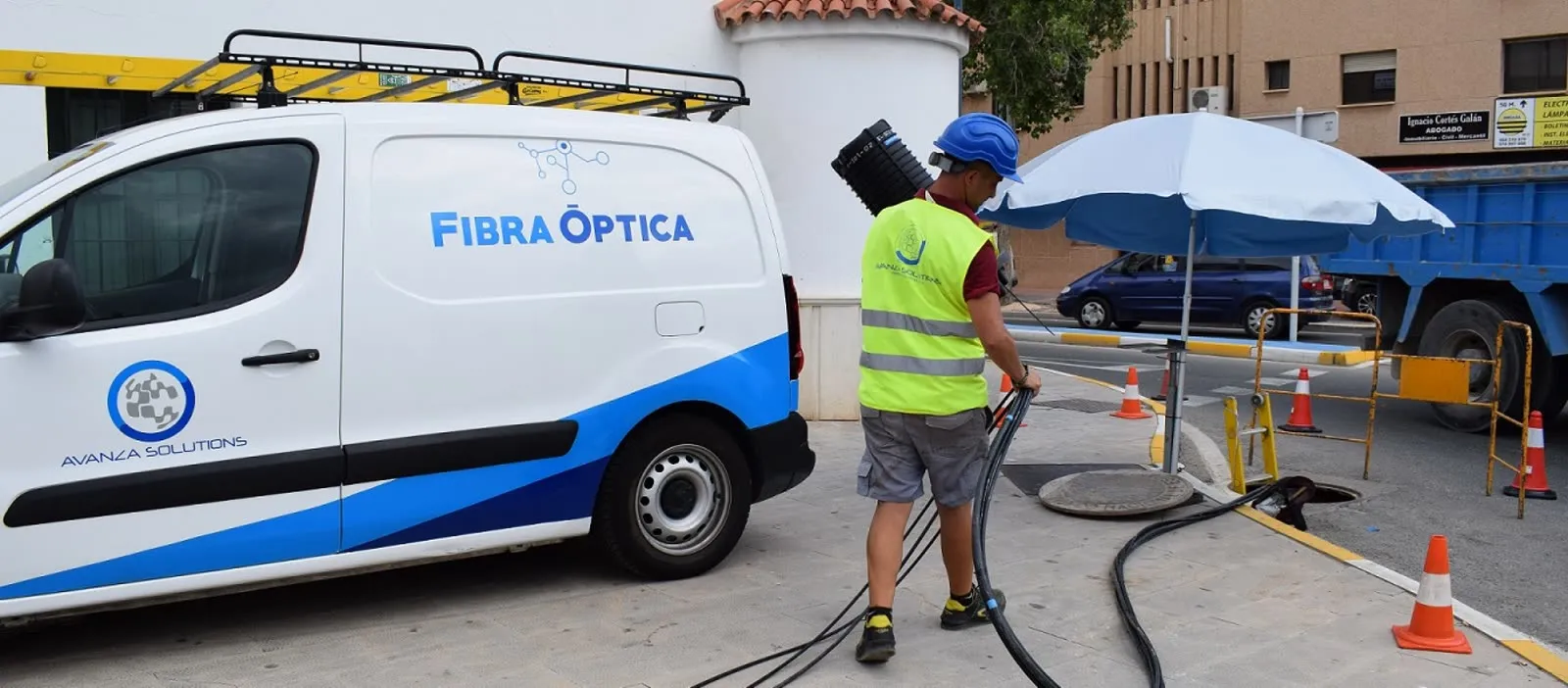 Operario con casco y chaleco reflectante manipula cable de fibra óptica junto a una furgoneta rotulada de Avanza Solutions y una arqueta abierta en la acera