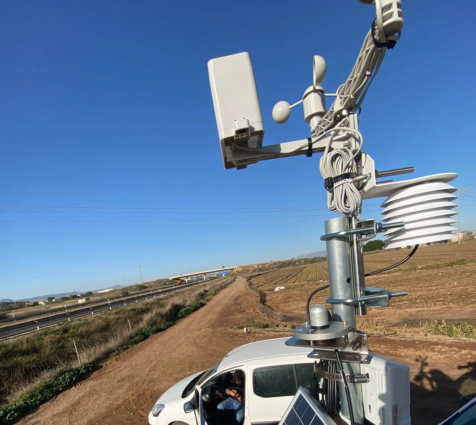 Mástil con sensores meteorológicos, módulo de comunicación y placa solar junto a un camino de tierra y una autovía